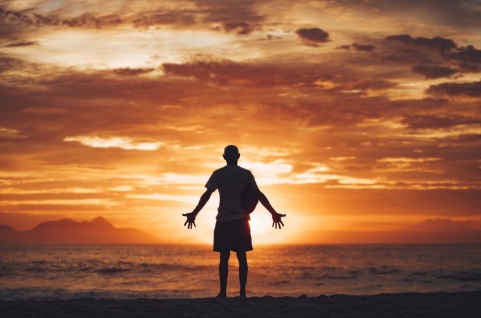 Imagem de silhueta de uma pessoa com os braços abertos apreciando a natureza no por do sol em frente ao mar