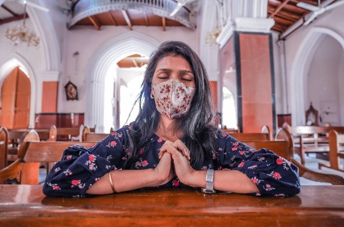 Imagem de uma jovem senhora ajoelhada, orando com os olhos fechados e de máscara, dentro de uma igreja vazia