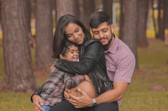 Imagem de uma mãe grávida sendo abraçada pelo filho e pelo pai com feição de felicidade e tranquilidade